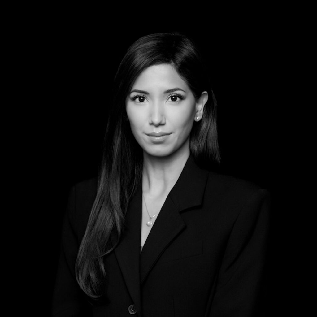 A woman with long straight hair is wearing a dark blazer and a necklace, looking directly at the camera with a neutral expression. The black-and-white image has a solid black background. Unified LLP | Toronto T. 416.787.7678 | Kitchener T. 519.729.5038 | Calgary T. 403-906-2351 | unifiedllp.com