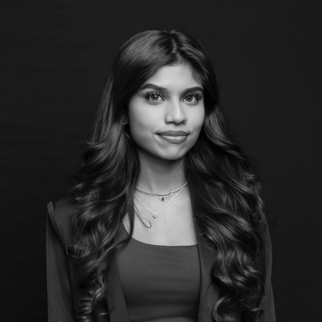 A young woman with long, wavy hair wears a jacket over a top and layered necklaces. She is posed against a dark background and looks directly at the camera with a neutral expression. The photo is in black and white. Unified LLP | Toronto T. 416.787.7678 | Kitchener T. 519.729.5038 | Calgary T. 403-906-2351 | unifiedllp.com