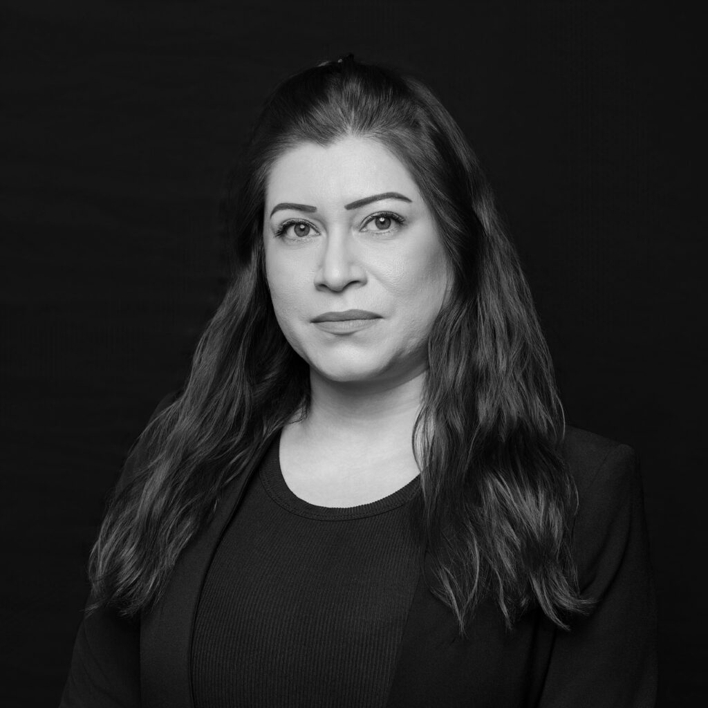 A woman with long, wavy hair and a calm expression is wearing a dark blazer and top. She is posed against a plain dark background in this black and white portrait. Unified LLP | Toronto T. 416.787.7678 | Kitchener T. 519.729.5038 | Calgary T. 403-906-2351 | unifiedllp.com