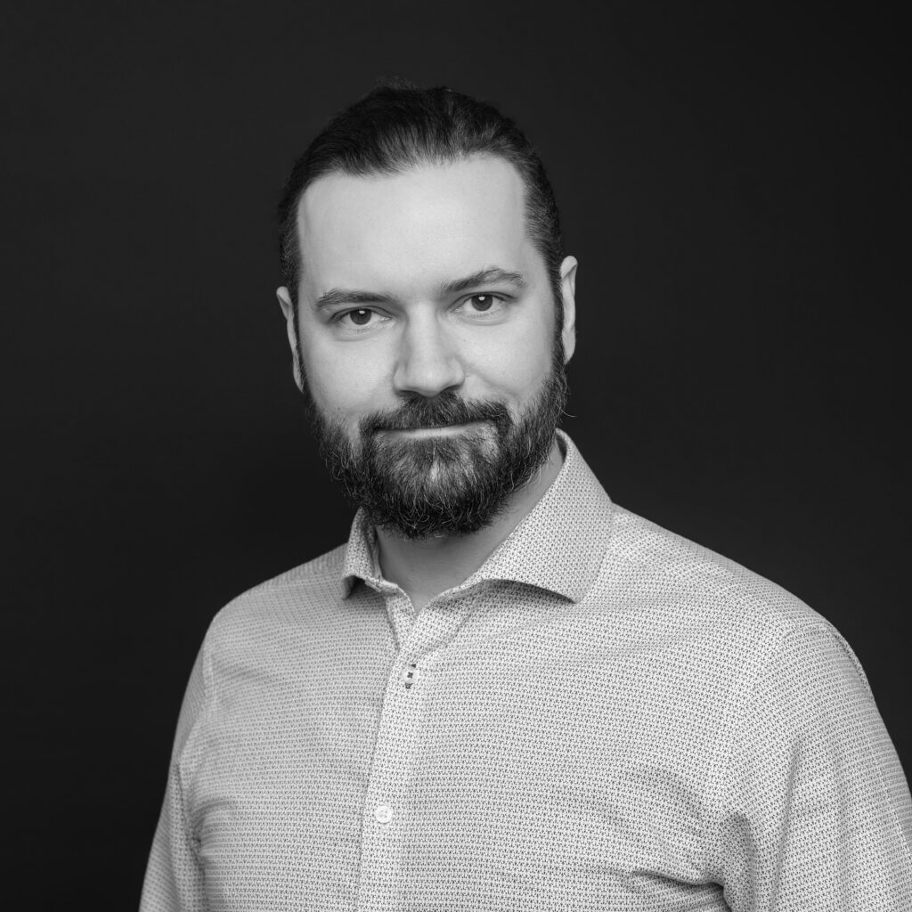 A man with a medium-length dark beard and slicked-back hair, wearing a light-coloured, patterned button-up shirt, poses against a plain dark background. The image is in black and white. Unified LLP | Toronto T. 416.787.7678 | Kitchener T. 519.729.5038 | Calgary T. 403-906-2351 | unifiedllp.com