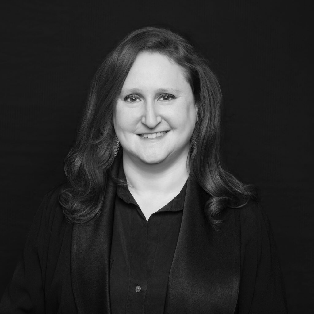 A woman with long wavy hair is smiling at the camera. She is wearing a dark blouse and a scarf, with a dark background behind her. The image is in black and white. Unified LLP | Toronto T. 416.787.7678 | Kitchener T. 519.729.5038 | Calgary T. 403-906-2351 | unifiedllp.com