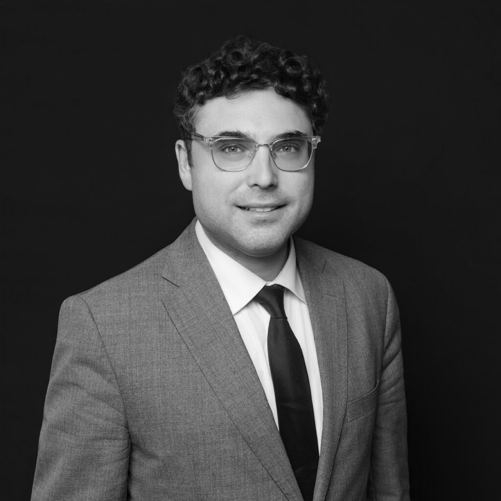 A man with short curly hair and glasses wearing a light-coloured suit, white shirt, and dark tie is posed against a plain dark background. He is facing forward and smiling slightly. Unified LLP | Toronto T. 416.787.7678 | Kitchener T. 519.729.5038 | Calgary T. 403-906-2351 | unifiedllp.com