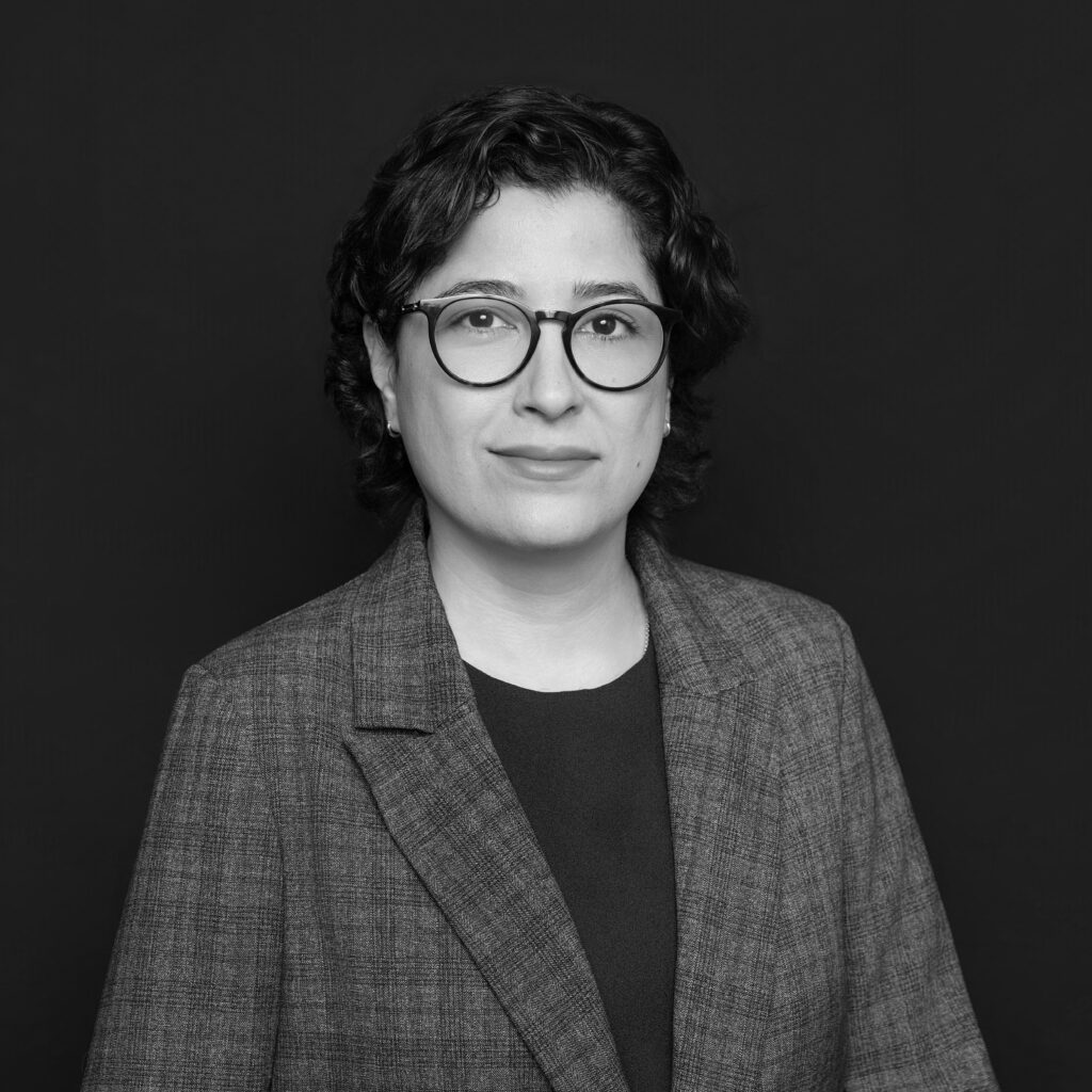 A person with short curly hair, wearing glasses, a checked blazer, and a dark top, poses for a formal portrait against a plain dark background. Unified LLP | Toronto T. 416.787.7678 | Kitchener T. 519.729.5038 | Calgary T. 403-906-2351 | unifiedllp.com
