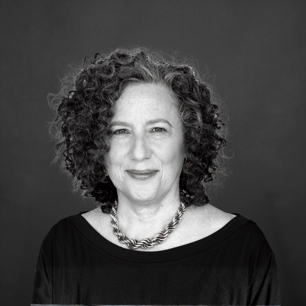 A middle-aged person with curly, greying hair and light skin is wearing a dark top and a twisted chain necklace, facing the camera and smiling slightly. The background is plain and dark. The image is in black and white. Unified LLP | Toronto T. 416.787.7678 | Kitchener T. 519.729.5038 | Calgary T. 403-906-2351 | unifiedllp.com