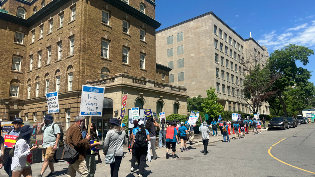 A group of people are marching on a city pavement holding signs with messages like Respect, Safety, Housing and Fair Wages Now during a protest on a sunny day. Old buildings and trees are visible in the background. Unified LLP | Toronto T. 416.787.7678 | Kitchener T. 519.729.5038 | Calgary T. 403-906-2351 | unifiedllp.com