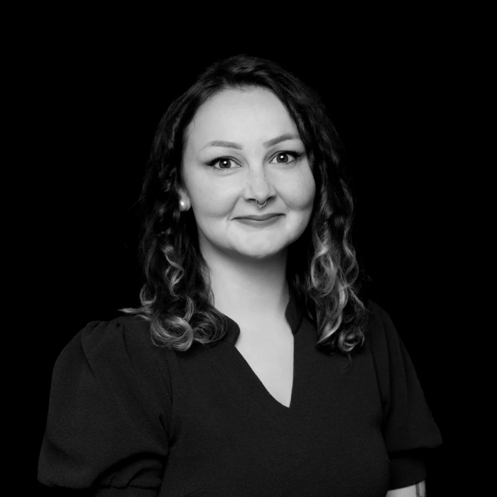 Black-and-white portrait of a person with wavy, shoulder-length hair, wearing a dark top. The person is facing forward, smiling slightly, and has a nose ring. The background is solid black. Unified LLP | Toronto T. 416.787.7678 | Kitchener T. 519.729.5038 | Calgary T. 403-906-2351 | unifiedllp.com
