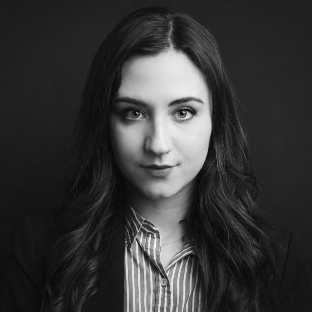 A woman with long, wavy hair wearing a pinstriped shirt and blazer stares directly at the camera with a neutral expression against a dark background. The photo is in black and white. Unified LLP | Toronto T. 416.787.7678 | Kitchener T. 519.729.5038 | Calgary T. 403-906-2351 | unifiedllp.com
