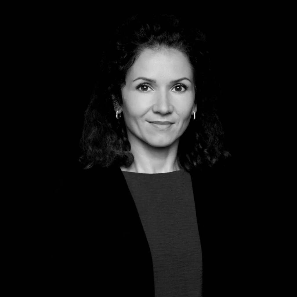 A woman with curly dark hair and hoop earrings, wearing a dark blazer over a light top, poses against a solid black background. She faces forwards with a slight smile, in a formal, professional portrait. Unified LLP | Toronto T. 416.787.7678 | Kitchener T. 519.729.5038 | Calgary T. 403-906-2351 | unifiedllp.com